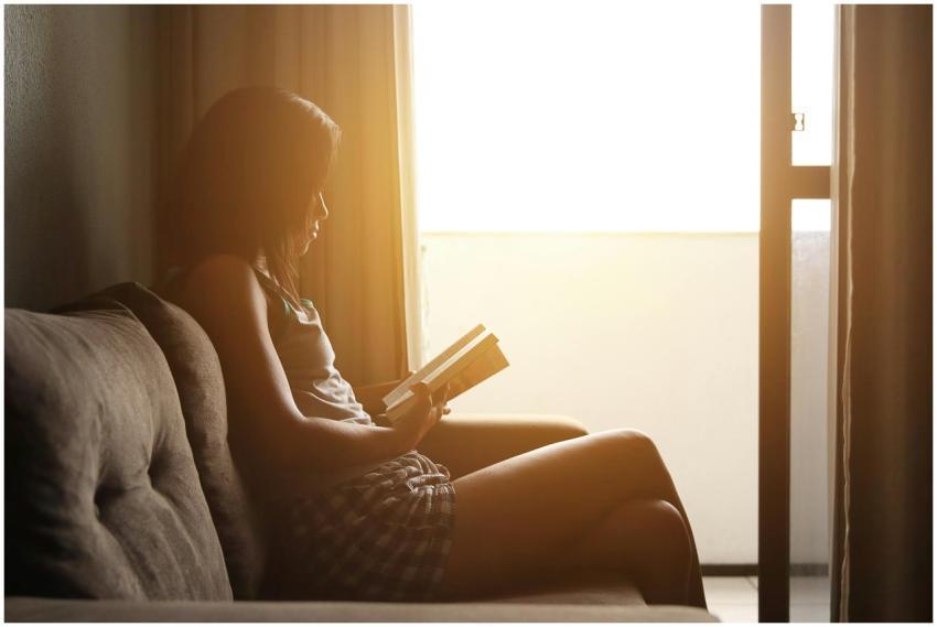 Woman reading a book on sofa by window with mornin