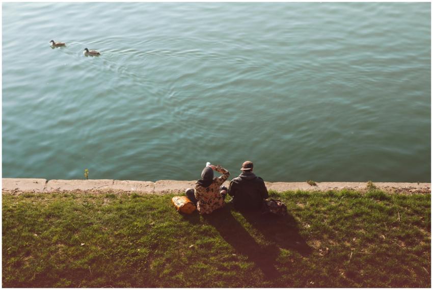 Two people sit by a serene lakeside watching ducks