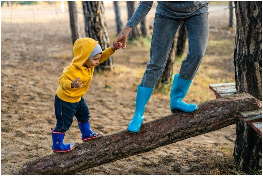 A woman helps her toddler walk on a log in a park,