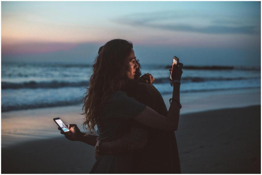 Couple embracing on a beach at sunset, using mobil