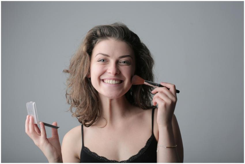 A joyful woman applies makeup with a brush, posing