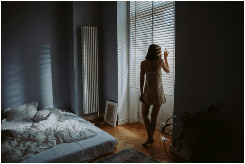 A woman in a bedroom stands by window blinds, cast