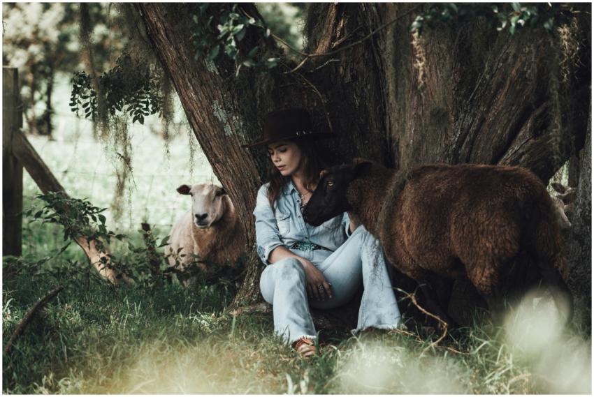 A woman in a jean jacket and hat sits with sheep u