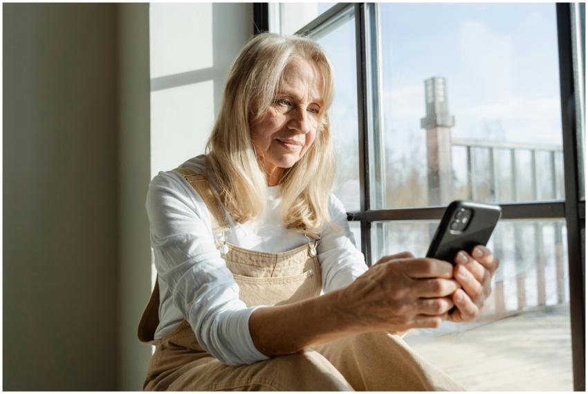 Elderly woman using a smartphone by a window, enjo