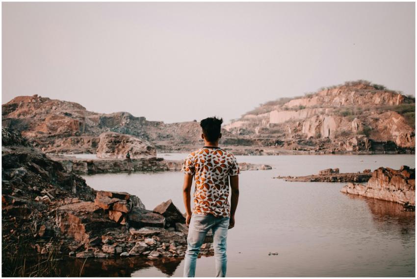 A man in a patterned shirt stands by a rocky lake,