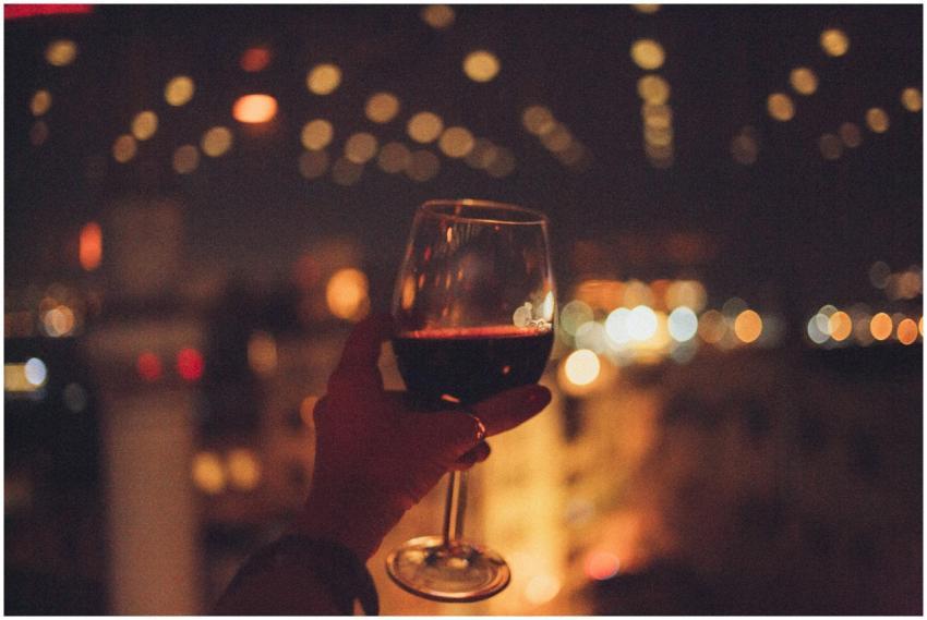 A hand holds a red wine glass with bokeh city ligh