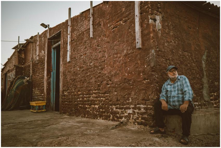 Elderly man sitting by a rustic building at sunset