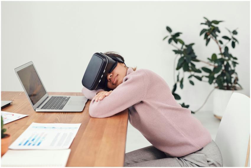 A woman wearing a VR headset rests on a desk besid