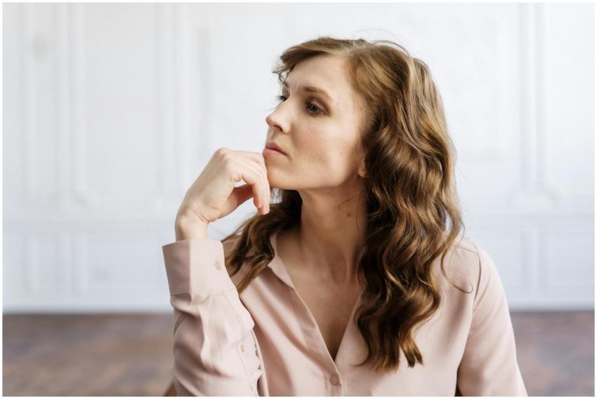 A thoughtful woman with curly hair sits indoors, r