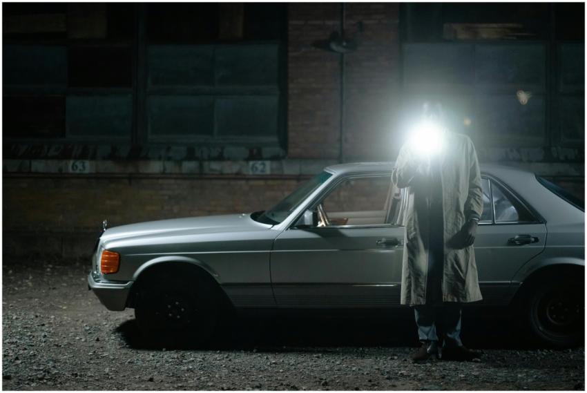 A detective stands by a vintage car at night, illu
