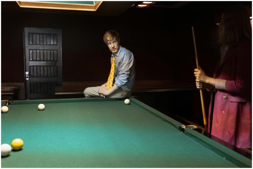 A man in a yellow tie sits on a billiard table whi