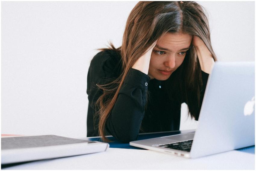 A stressed woman at a desk, looking at a laptop wi