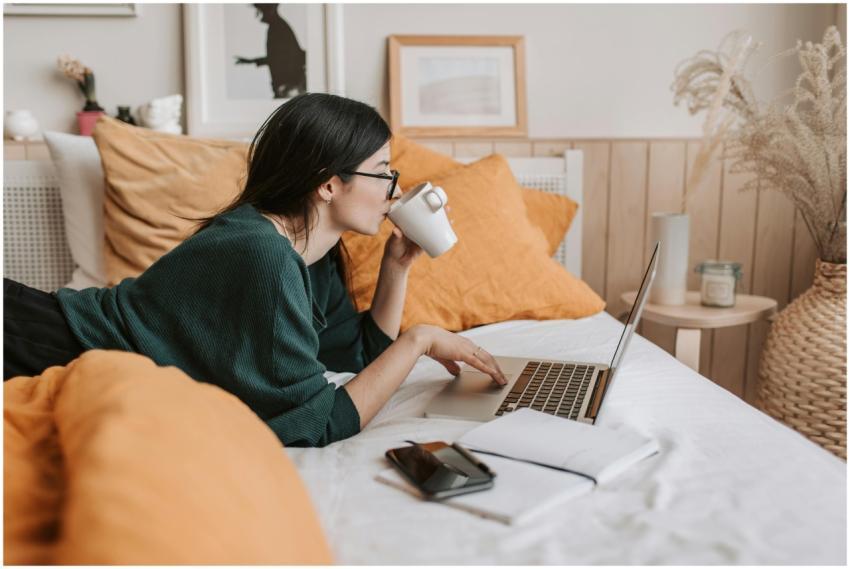 Woman relaxing on bed with coffee browsing laptop