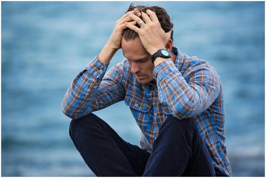 A man in a plaid shirt sits by the water looking d