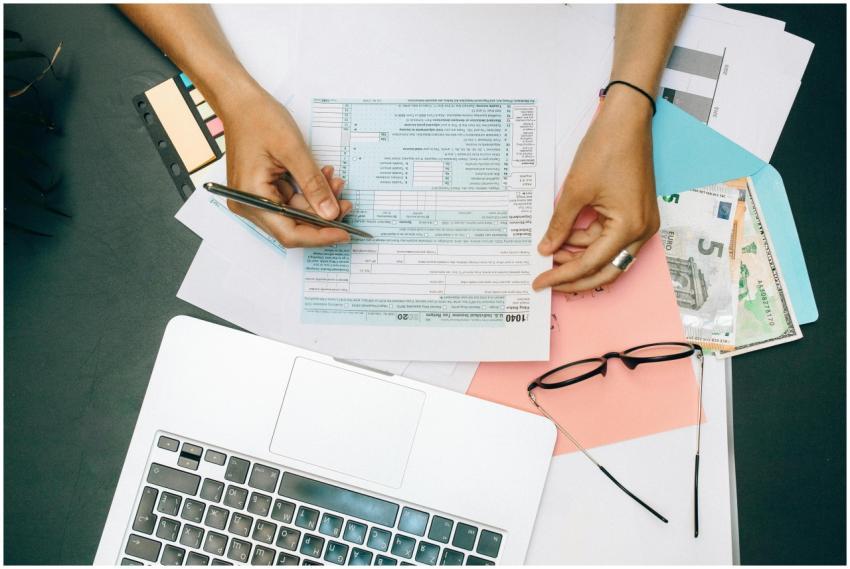 Hands writing on tax documents with laptop, glasse