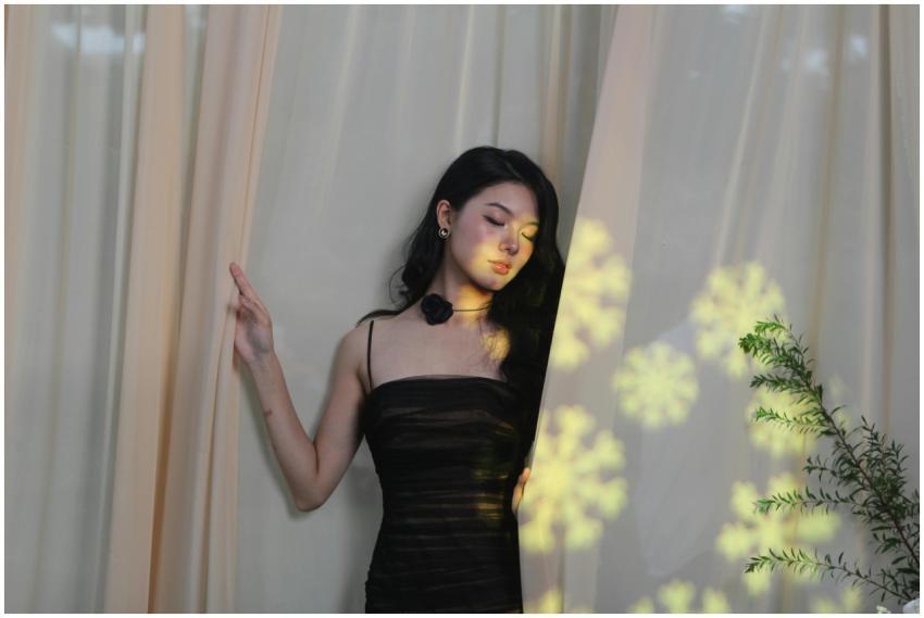 A woman in a black dress stands by curtains, with