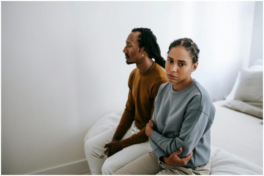 A couple sitting silently on a bed with tense expr