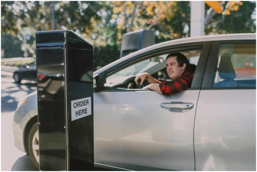 A man in a car orders at a drive-thru window, surr