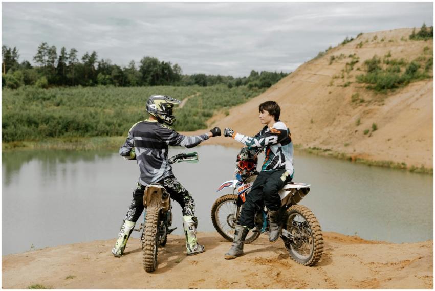 Two dirt bike riders fist bump at a scenic lakesid