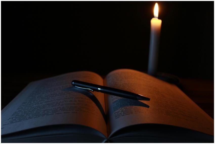 An open book and pen on a desk illuminated by cand