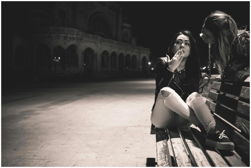 Two women converse on a city bench at night, expre