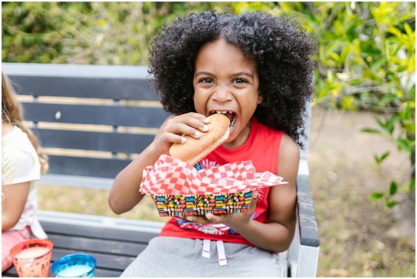 A joyful child with curly hair enjoys a hotdog on