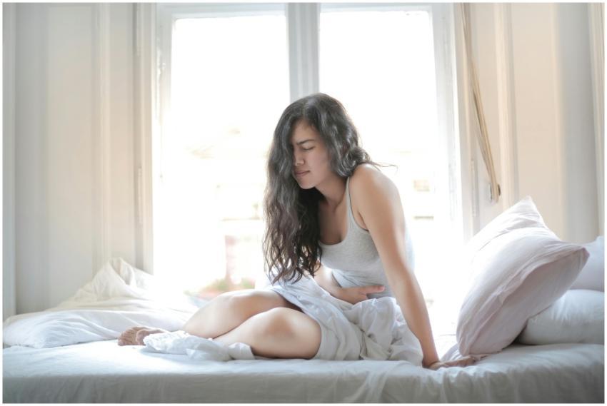 Woman sitting on bed holding her stomach, showing