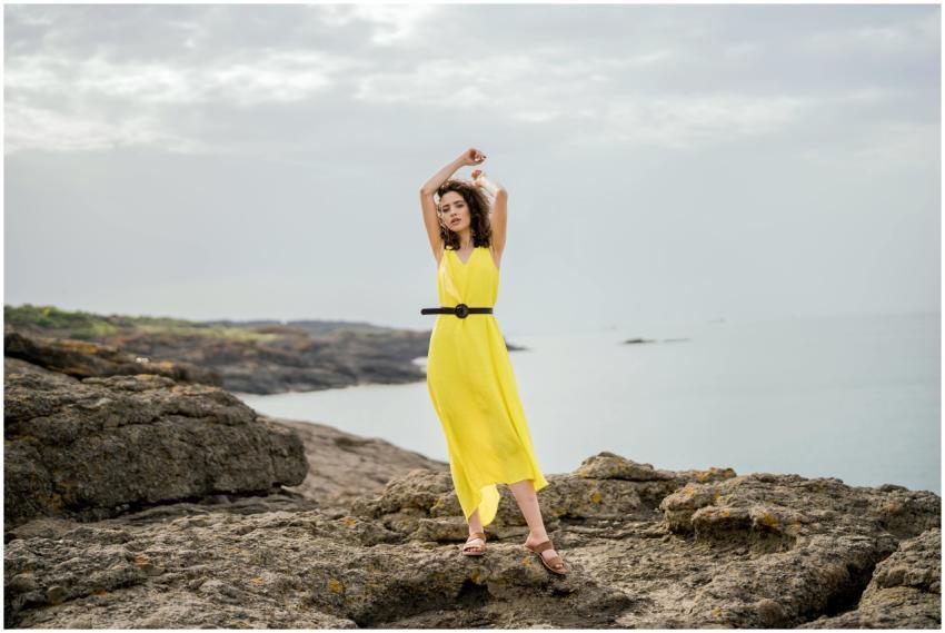 A woman in a yellow dress poses on rocky seaside t