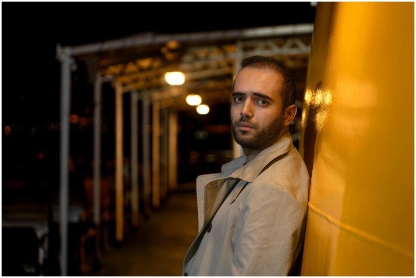 A man in a trench coat leans against a yellow wall