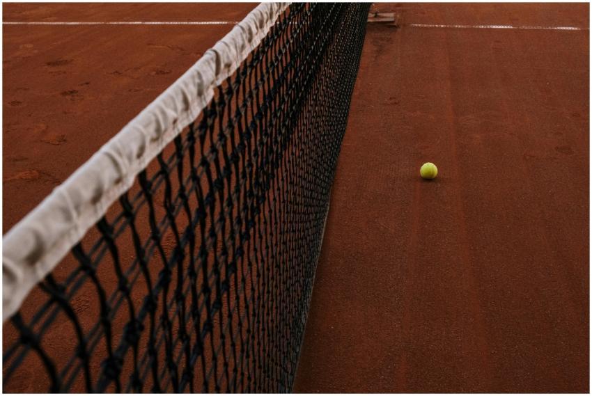 A tennis ball rests on a clay court next to the ne