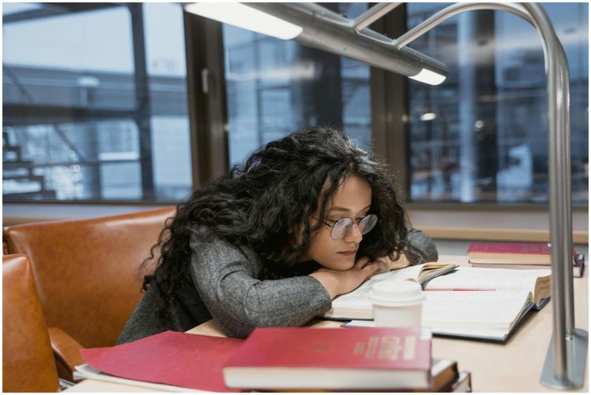 Tired young woman studying at a library desk, surr