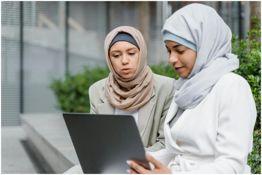 Two women in hijabs collaborate on a laptop outdoo