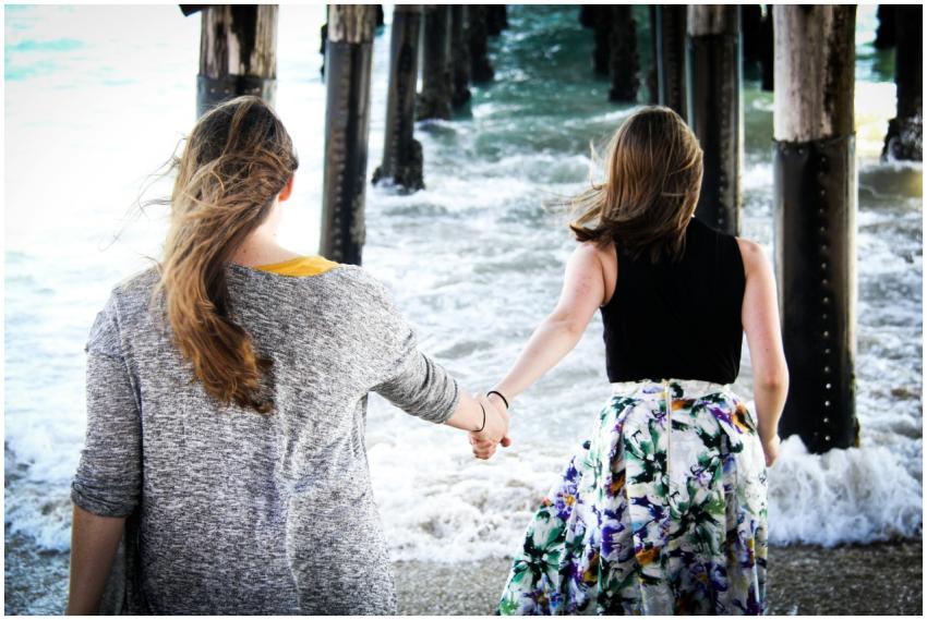 Two women holding hands under a pier by the ocean,