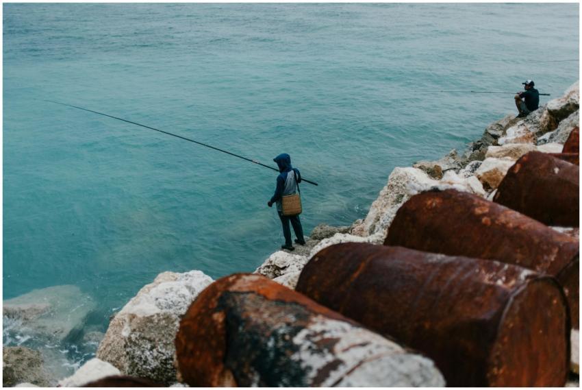 Two people fishing on a rocky shore by the sea, ca