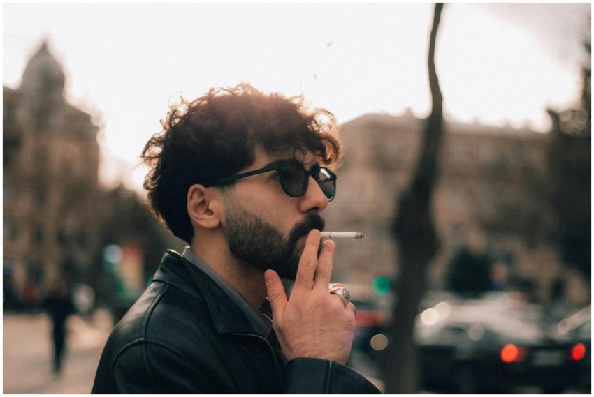 A man wearing sunglasses and a leather jacket smok