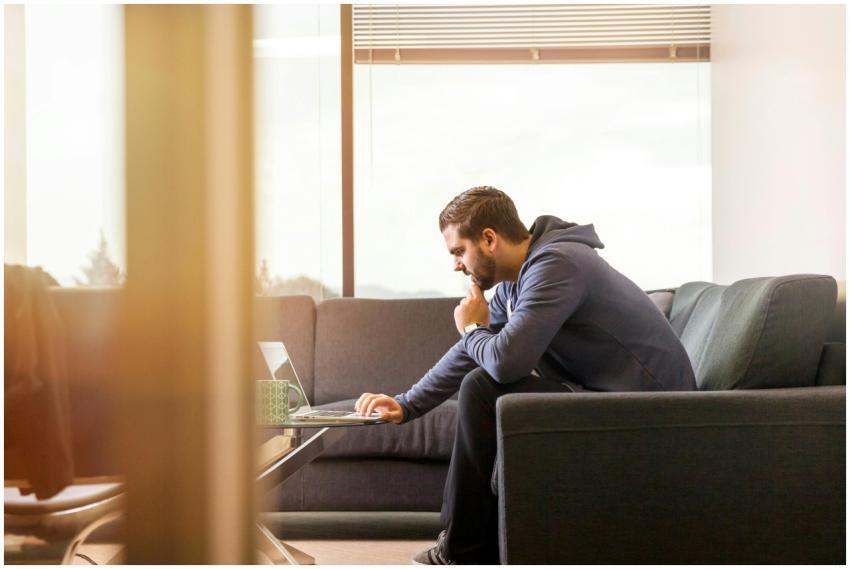 A man in casual attire works remotely from a sofa,