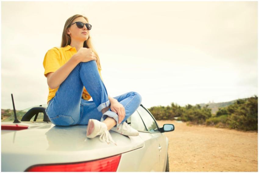 Young woman wearing sunglasses sits on car roof, e