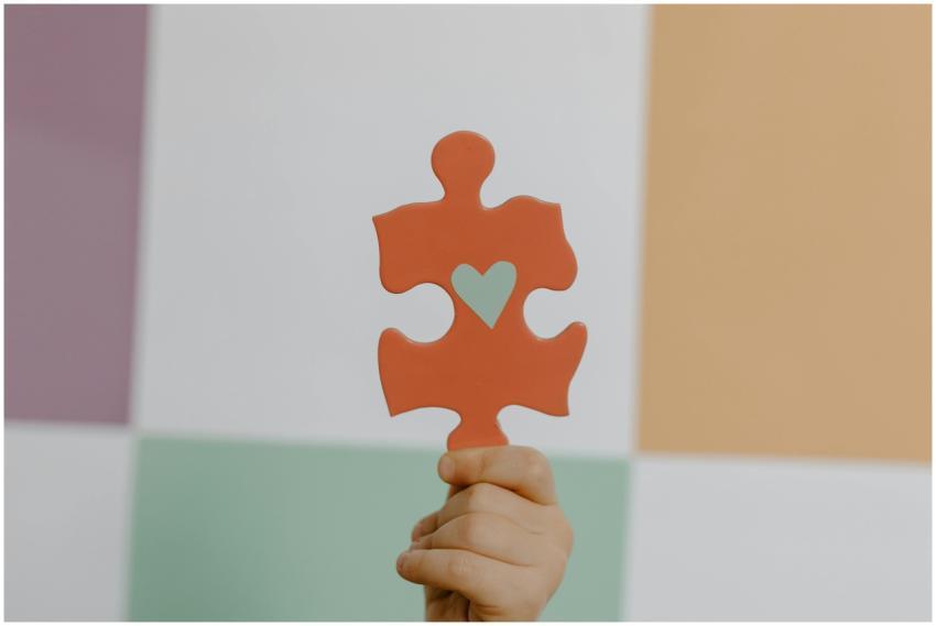 A hand holds a red puzzle piece with a heart, symb