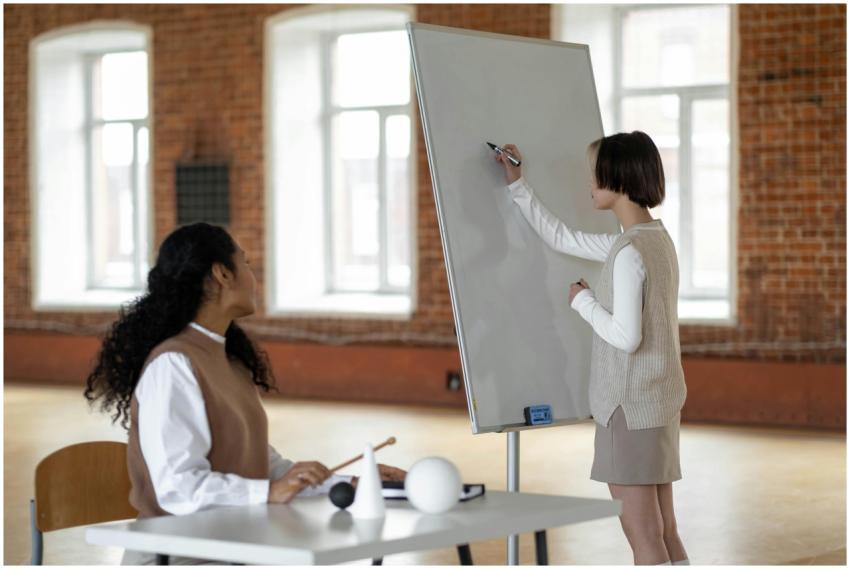 A student writes on a whiteboard watched by a teac