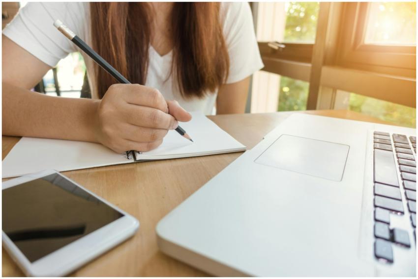 A young woman writing notes at a table with laptop