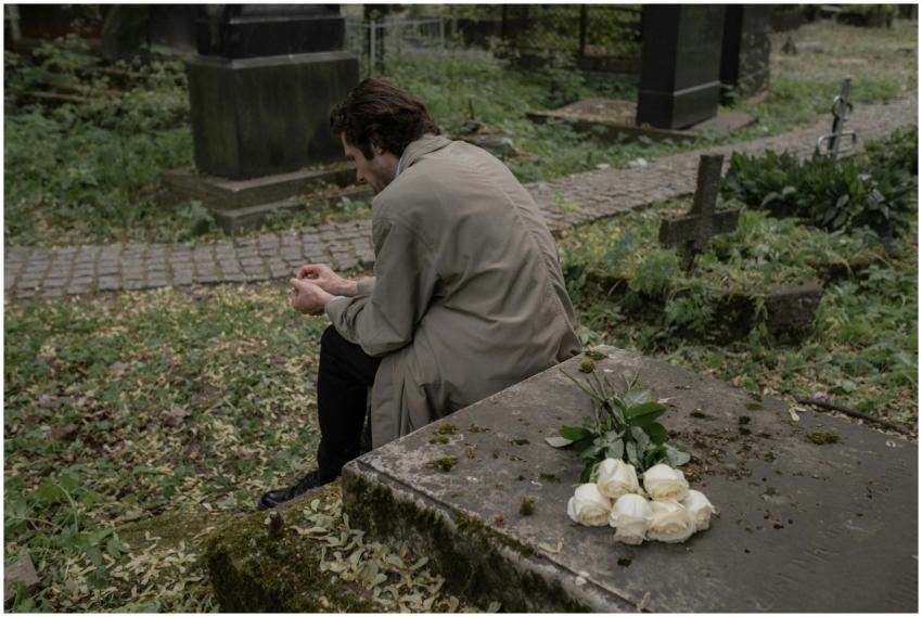 A man in a trench coat sits grieving at a cemetery