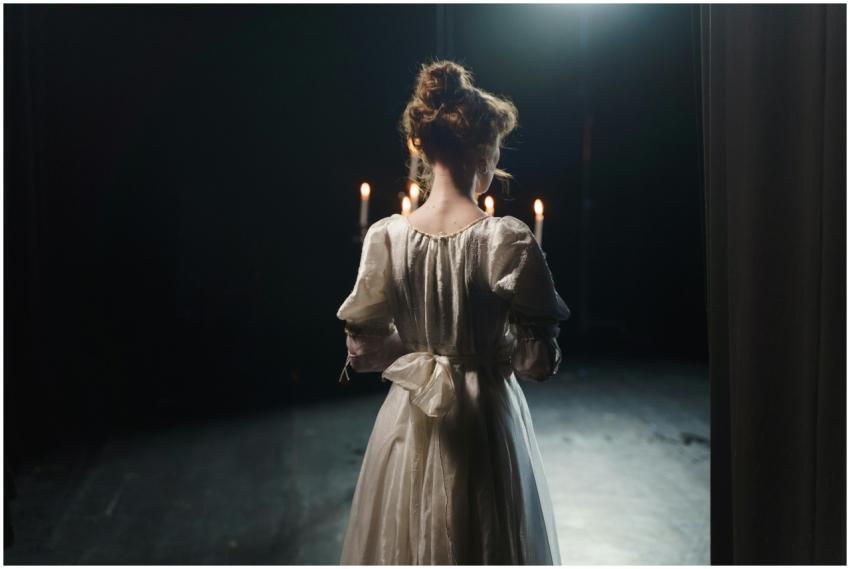 An actress in a white dress stands backstage in di