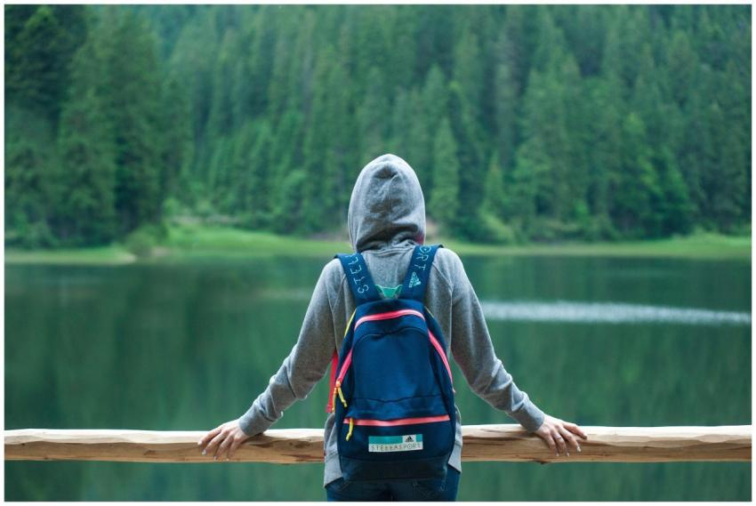 A person in a hoodie stands by a serene lake surro