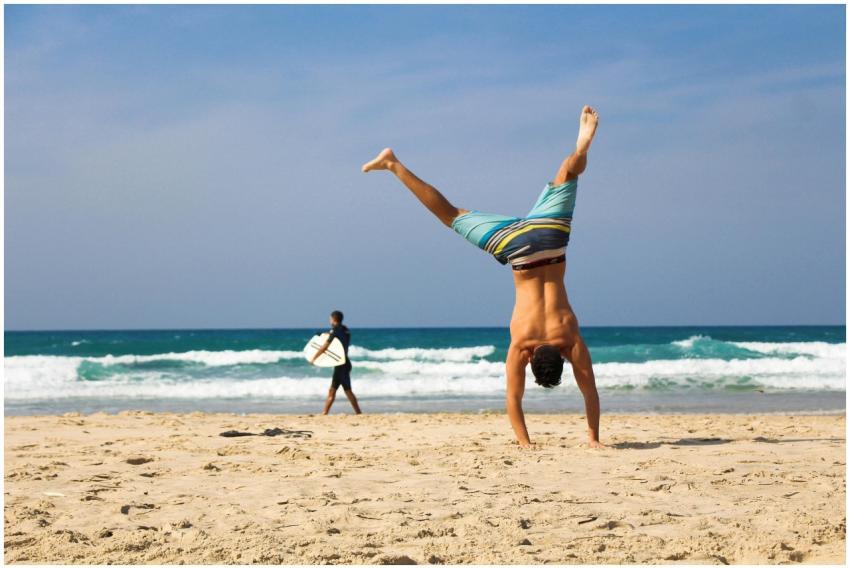 Man doing a handstand on a sunny beach with surfer