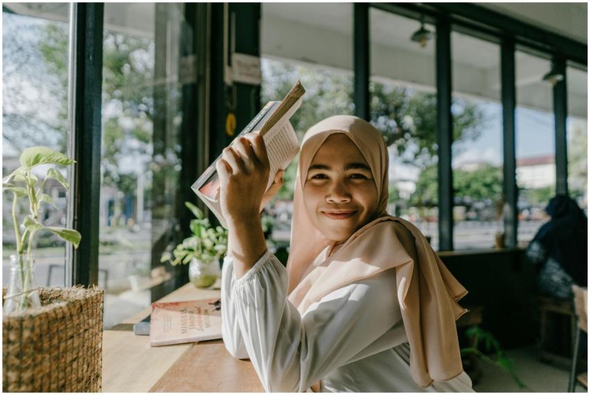 A smiling woman in a hijab reading a book by a win