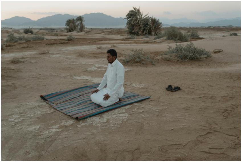 A man kneels in prayer on a mat in a desert landsc