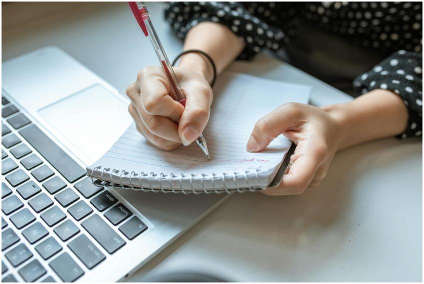 Person writing in a spiral notebook next to a lapt