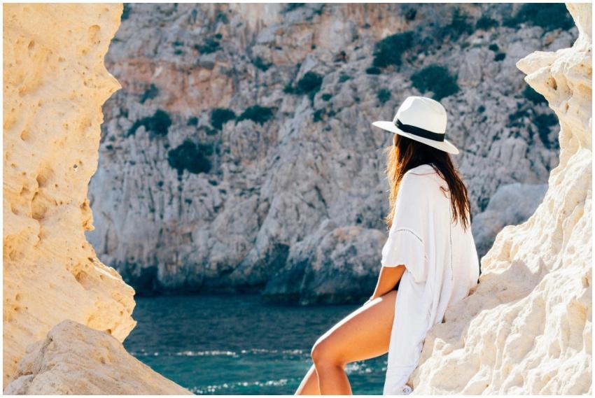 A woman in a hat sits on a rocky beach in summer,