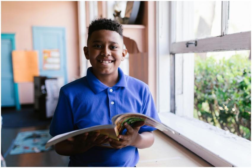 Young boy in blue polo reading by a classroom wind