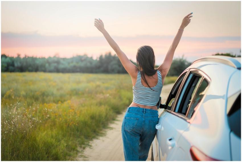 A woman stands beside a car on a summer field road